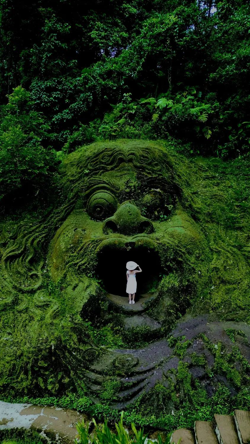 Moss-covered giant stone face carving with an open-mouth entrance in a lush jungle setting, with a person standing inside, in Bali, Indonesia.