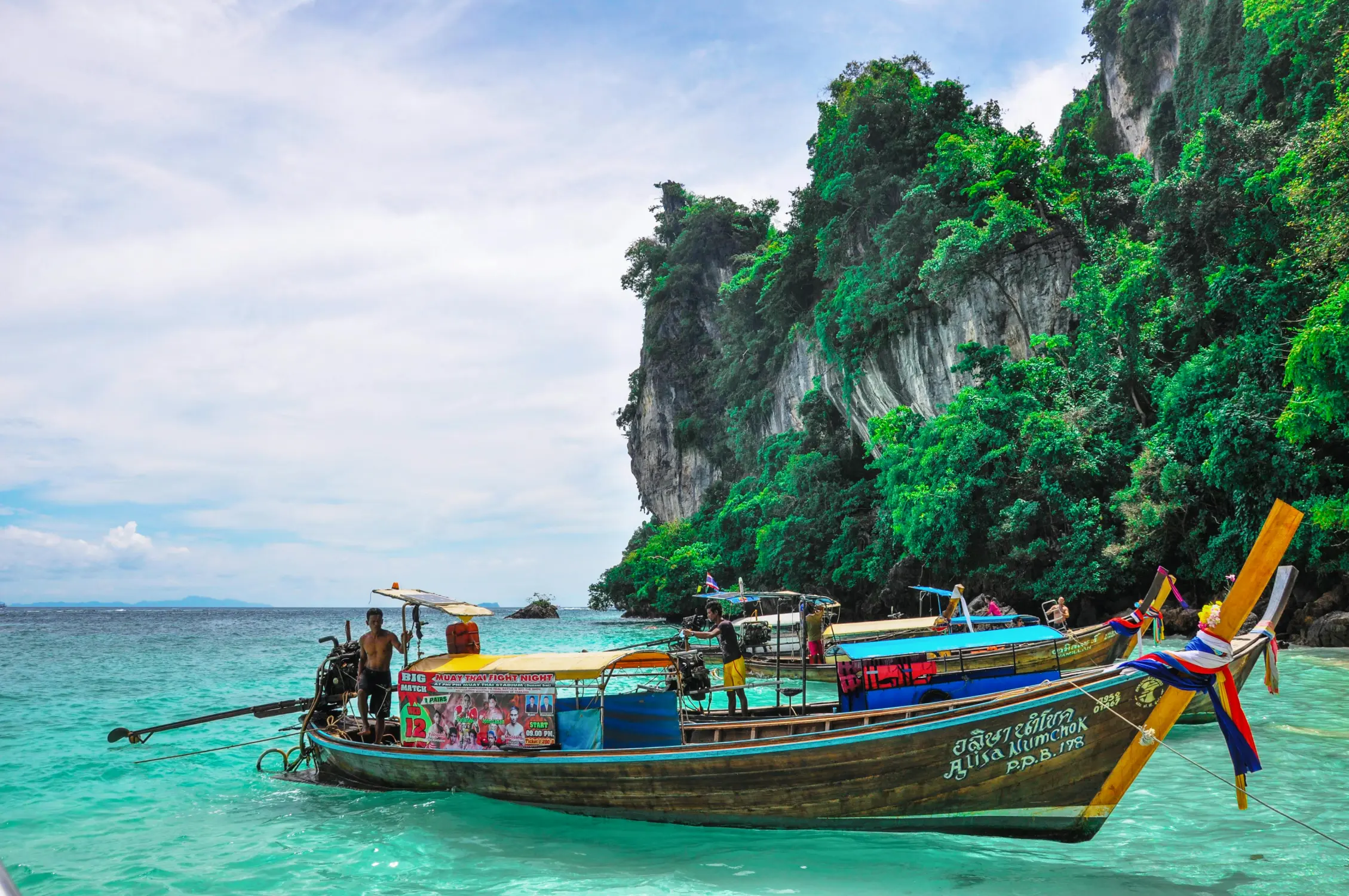 Colorful traditional Thai longtail boats with local boatmen anchored in crystal clear turquoise water against towering jungle-covered limestone karst cliffs at Koh Phi Phi, Krabi, Thailand