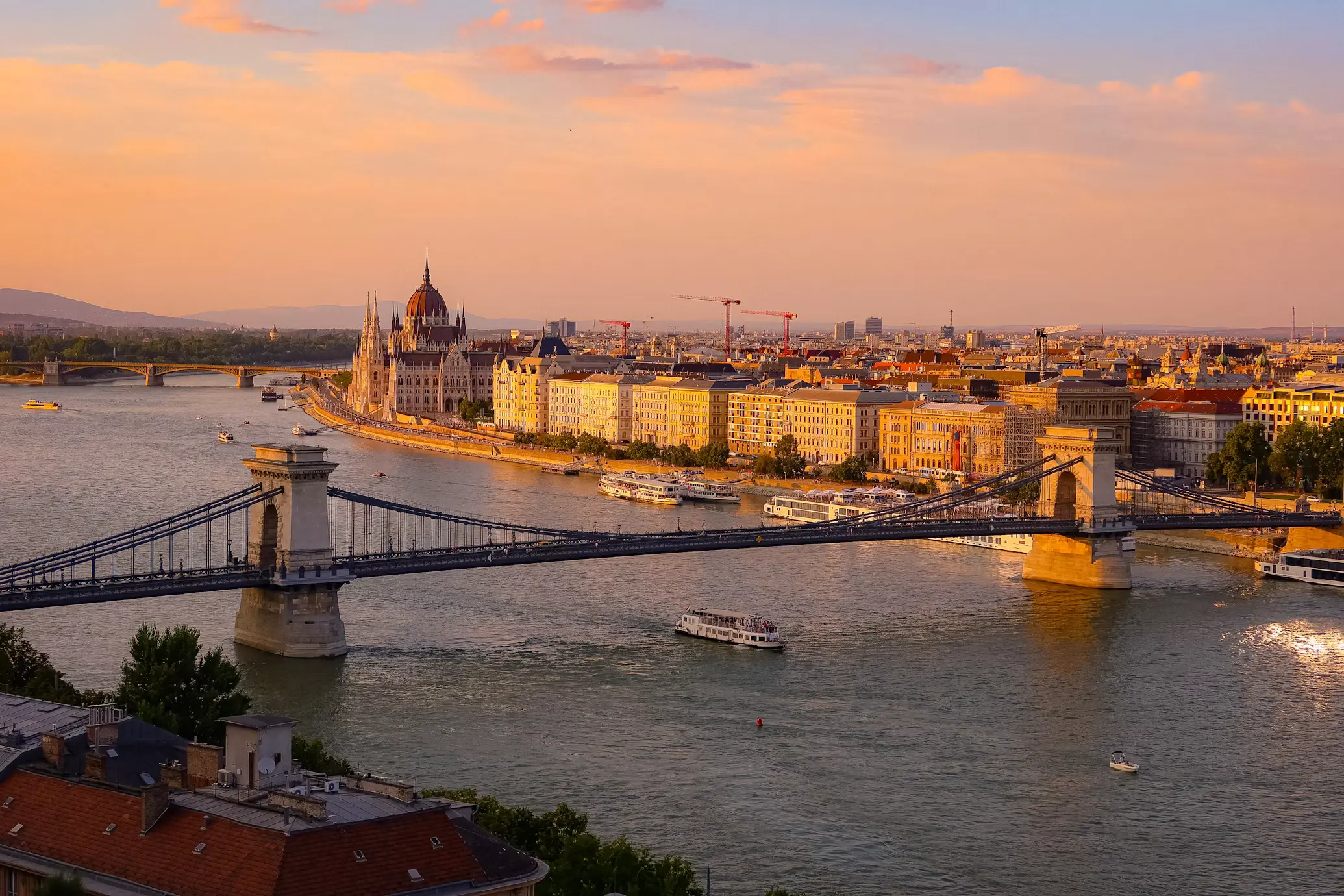 Suspension bridge spanning a wide river with a grand riverside parliament building and city skyline at sunset.
