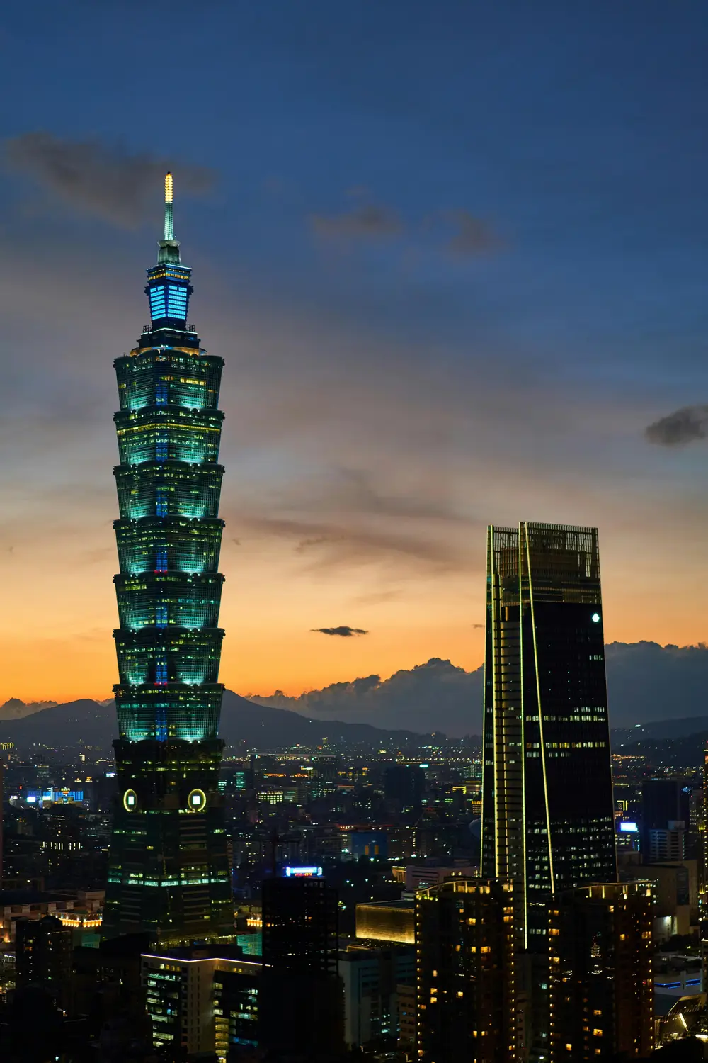 Taipei 101 skyscraper illuminated at twilight against an orange sunset sky with the Taipei city skyline and mountains
