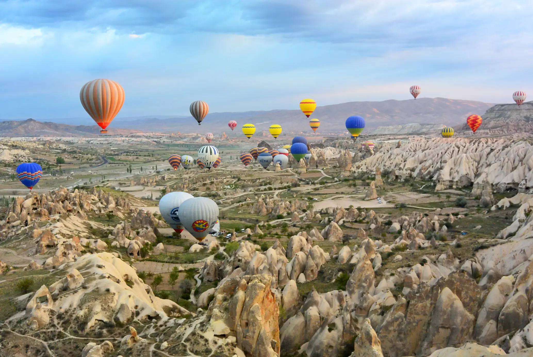 Dozens of colorful hot air balloons flying over the rocky landscape of Cappadocia, Turkey, with valleys and fairy chimney rock formations below.