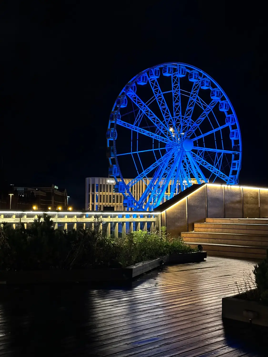 Blue-lit Ferris wheel at night beside a wooden deck with illuminated railings and steps.