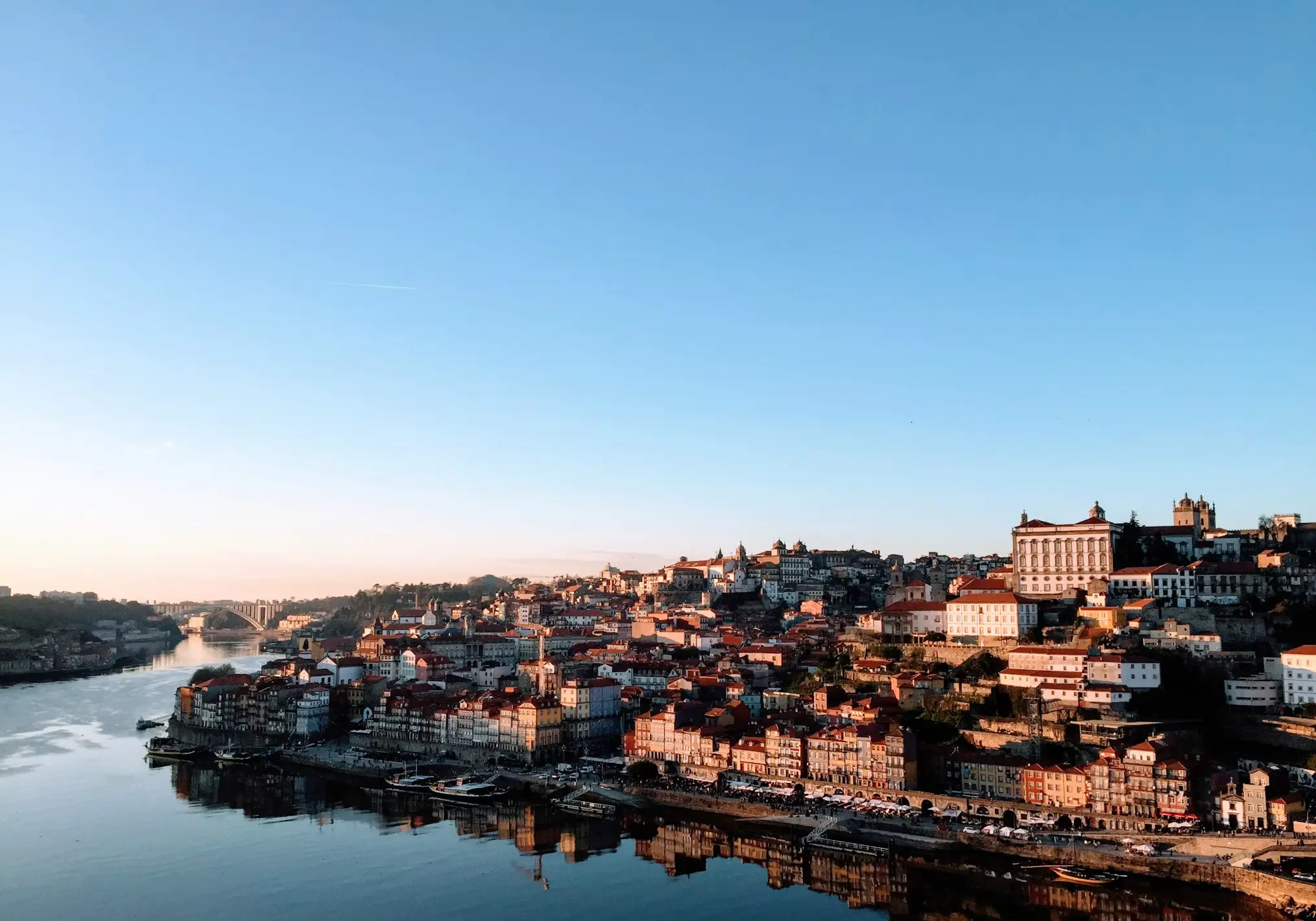 Colorful riverside city with historic buildings on a hillside, reflected in a calm river under a clear blue sky.
