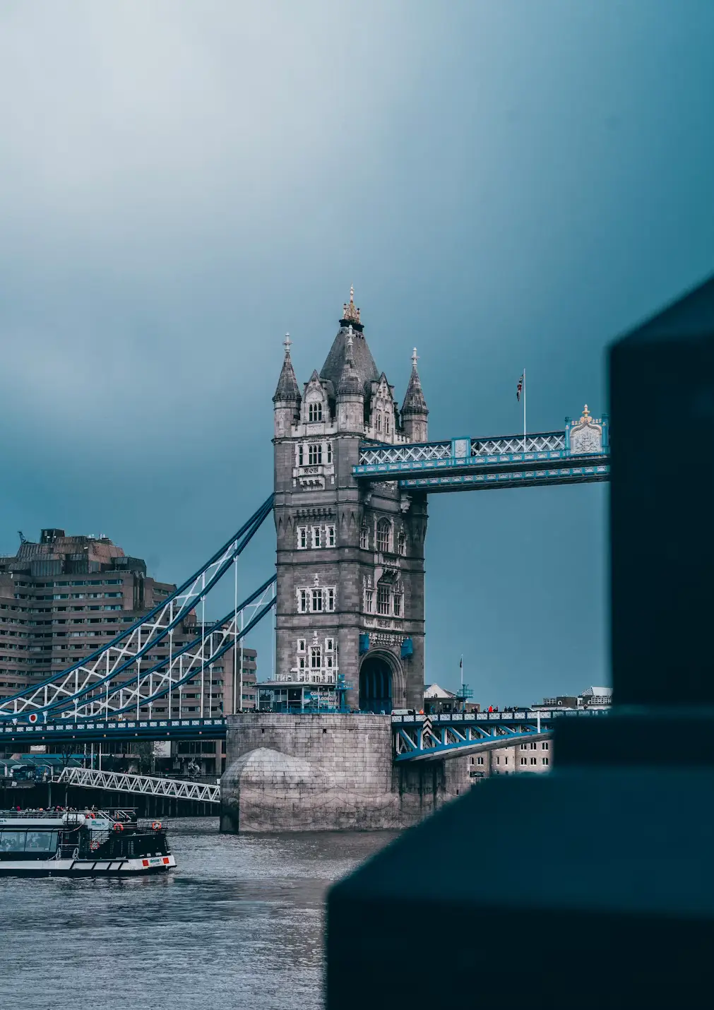 Moody view of Tower Bridge under an overcast sky, its stone towers and blue steelwork framed by a dark structure in the foreground.