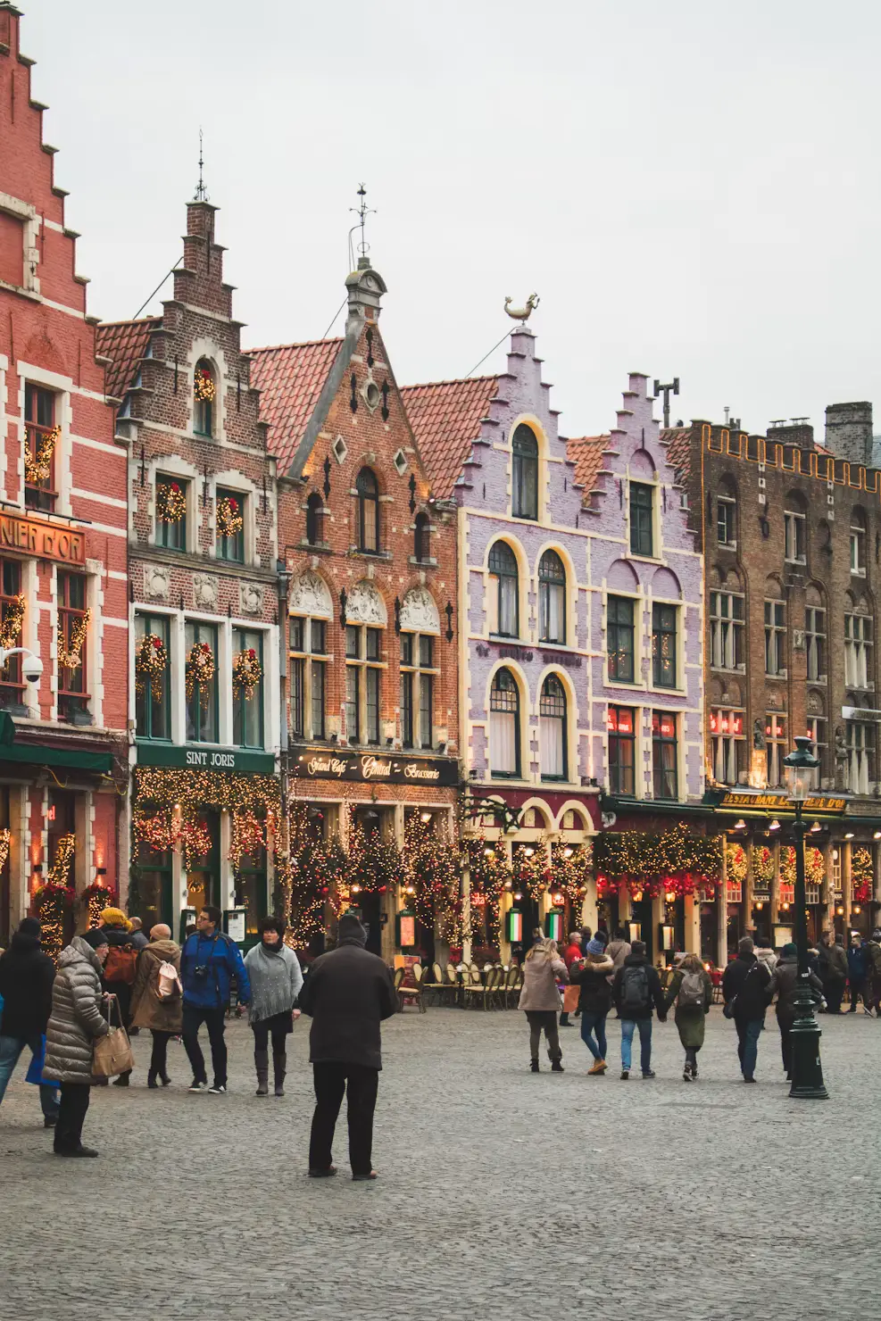 Colorful stepped-gable guild houses decorated with holiday lights around Bruges' Markt square, with people walking across the cobblestones.