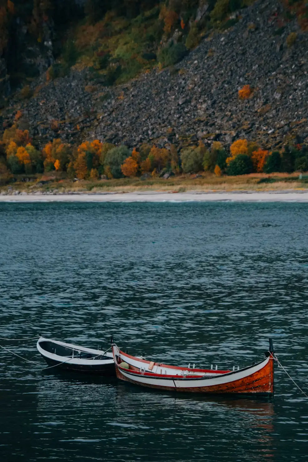 Two small wooden rowboats floating on dark rippled water, with a rocky slope and autumn trees on the far shore.