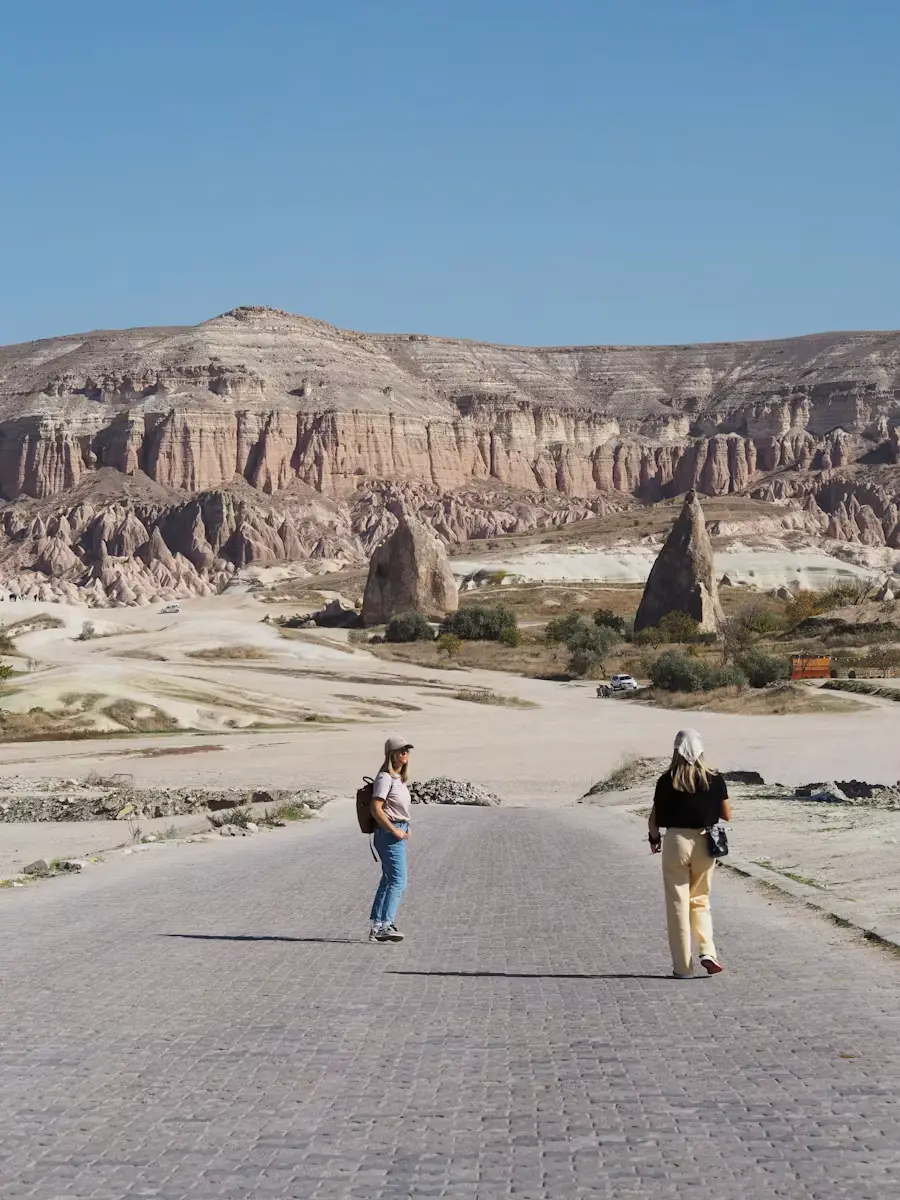Two people walking along a paved road through the Cappadocia valley, surrounded by unique rock formations and cave structures.