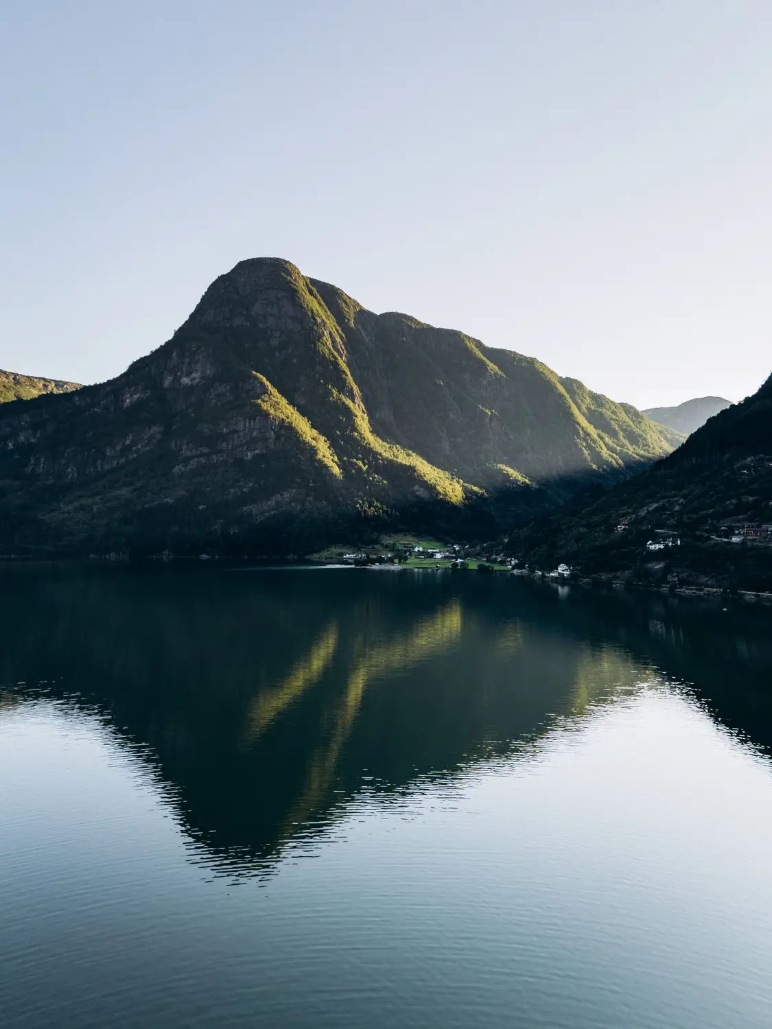 A sunlit mountain peak mirrored perfectly in the still waters of a fjord at dawn.