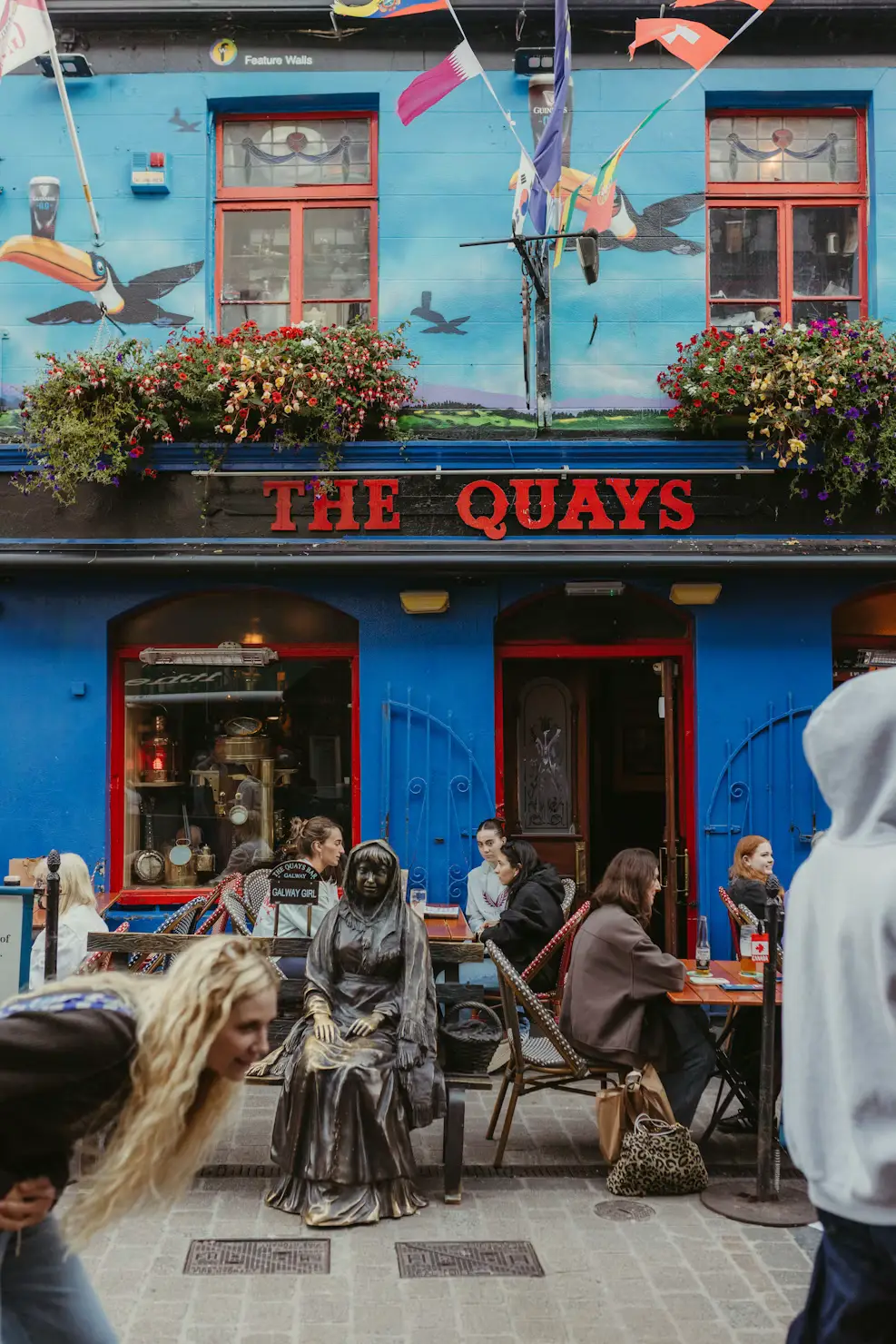 Blue pub building called 'The Quays' with murals and flags, people seated at outdoor tables.