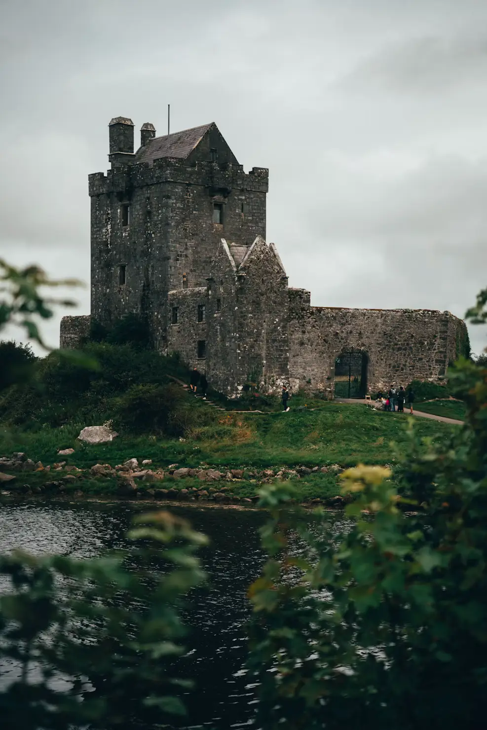 Stone tower castle beside a lake, surrounded by greenery under an overcast sky, Ireland.