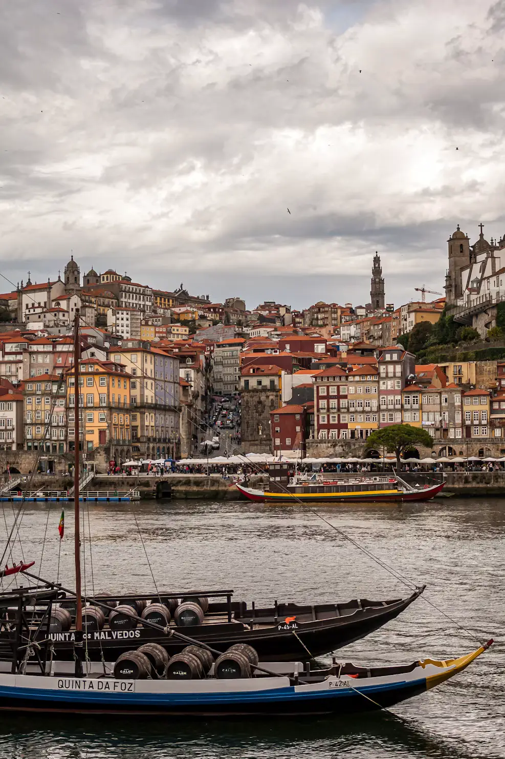 Traditional boats on a river with a hillside of colorful buildings and church towers under cloudy skies.