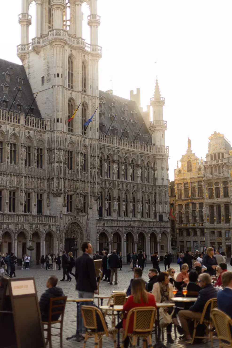 Sunlit Gothic facade of Brussels' Town Hall with outdoor cafe tables and visitors in the Grand Place.