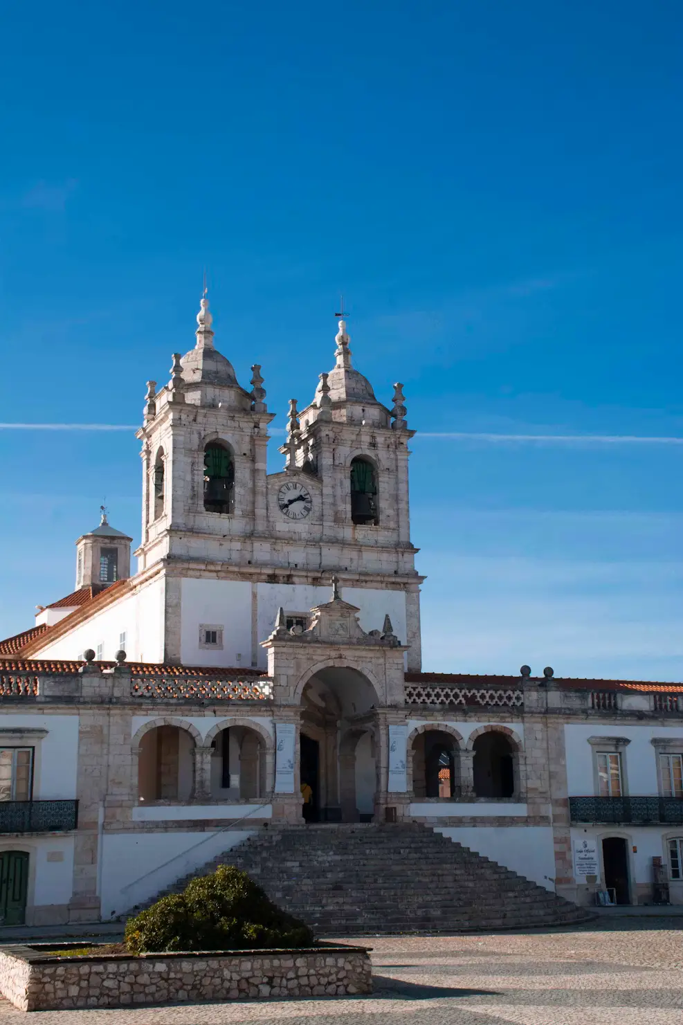 A white church with twin bell towers, a central clock, and stone steps beneath a clear blue sky.