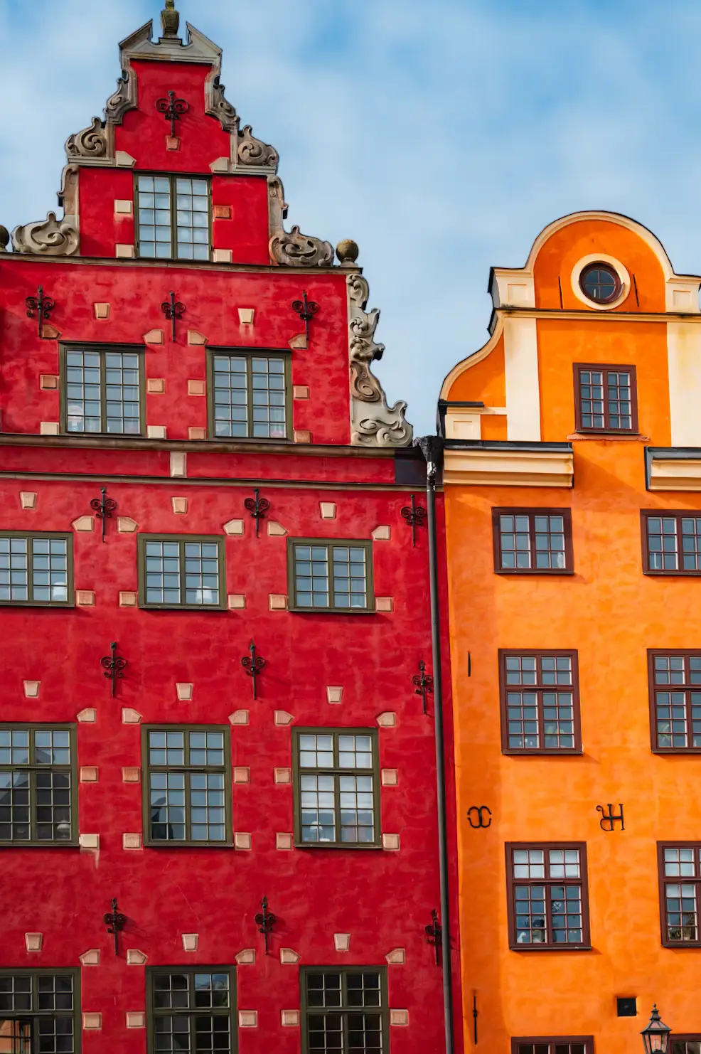 A tall, deep red stepped-gable building with ornate detailing stands beside an orange building, set against a bright blue sky.