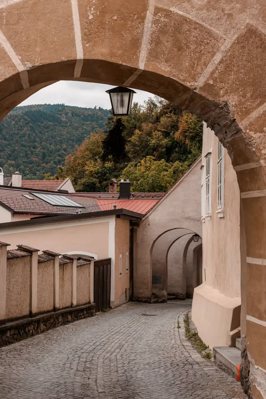 A stone archway framing a view of a quiet cobblestone lane leading toward green hills in the distance.
