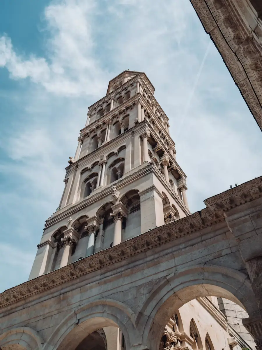 Low-angle view of a tall stone bell tower with arched windows, rising above a colonnaded building against a partly cloudy sky.