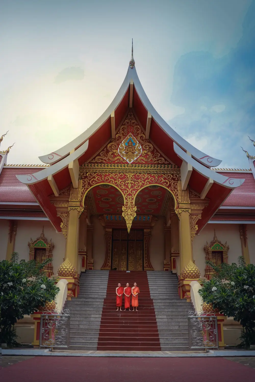 Ornate red and gold Buddhist temple with monks in robes on its steps in Luang Prabang, Laos.