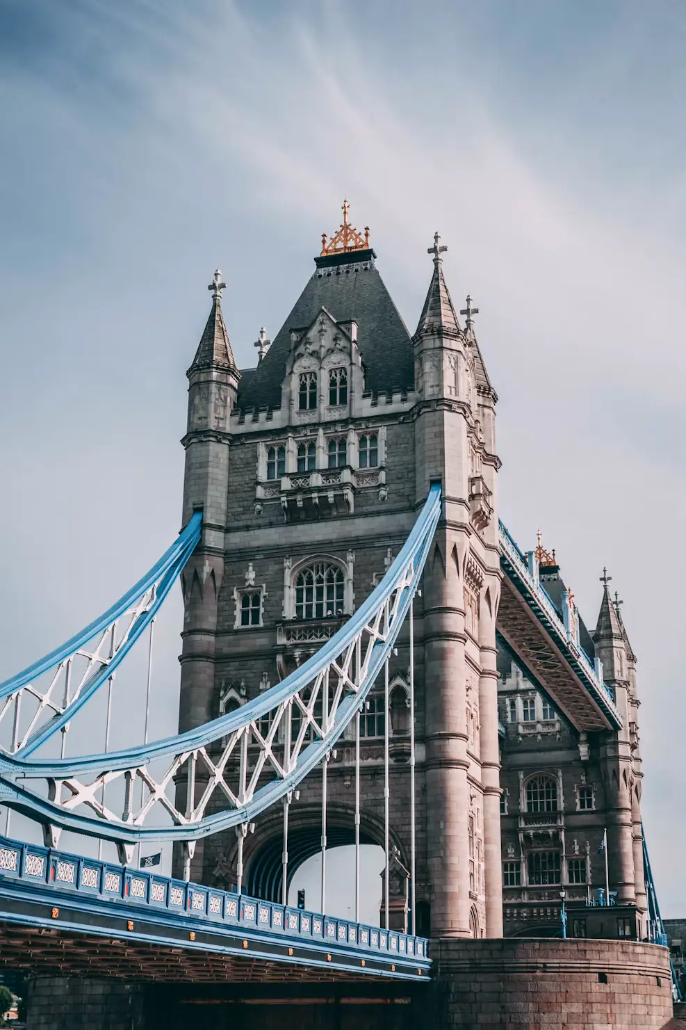 A close-up view of one of Tower Bridge's Gothic stone towers with its blue steel suspension supports, set against a soft sky.