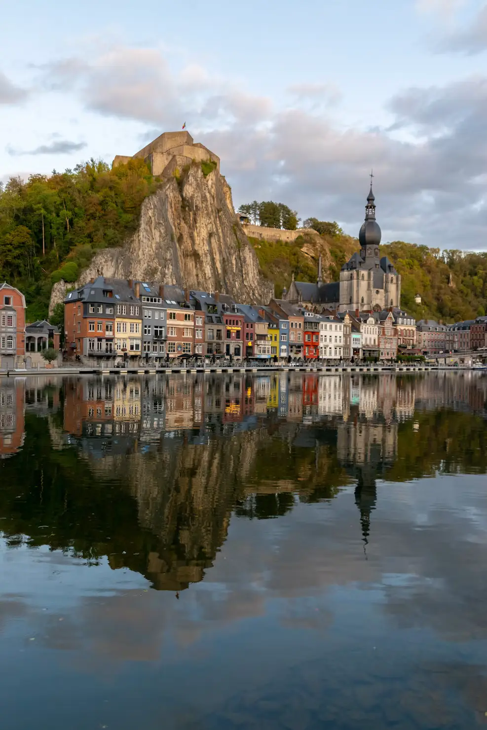The colorful riverside houses of Dinant beneath a steep cliff and citadel, with the onion-domed church tower reflected in the Meuse River.