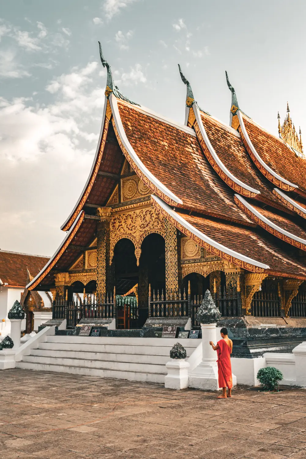Buddhist monk in orange robes standing before the ornate gold-decorated Wat Xieng Thong temple with its sweeping tiered roof in Luang Prabang, Laos.