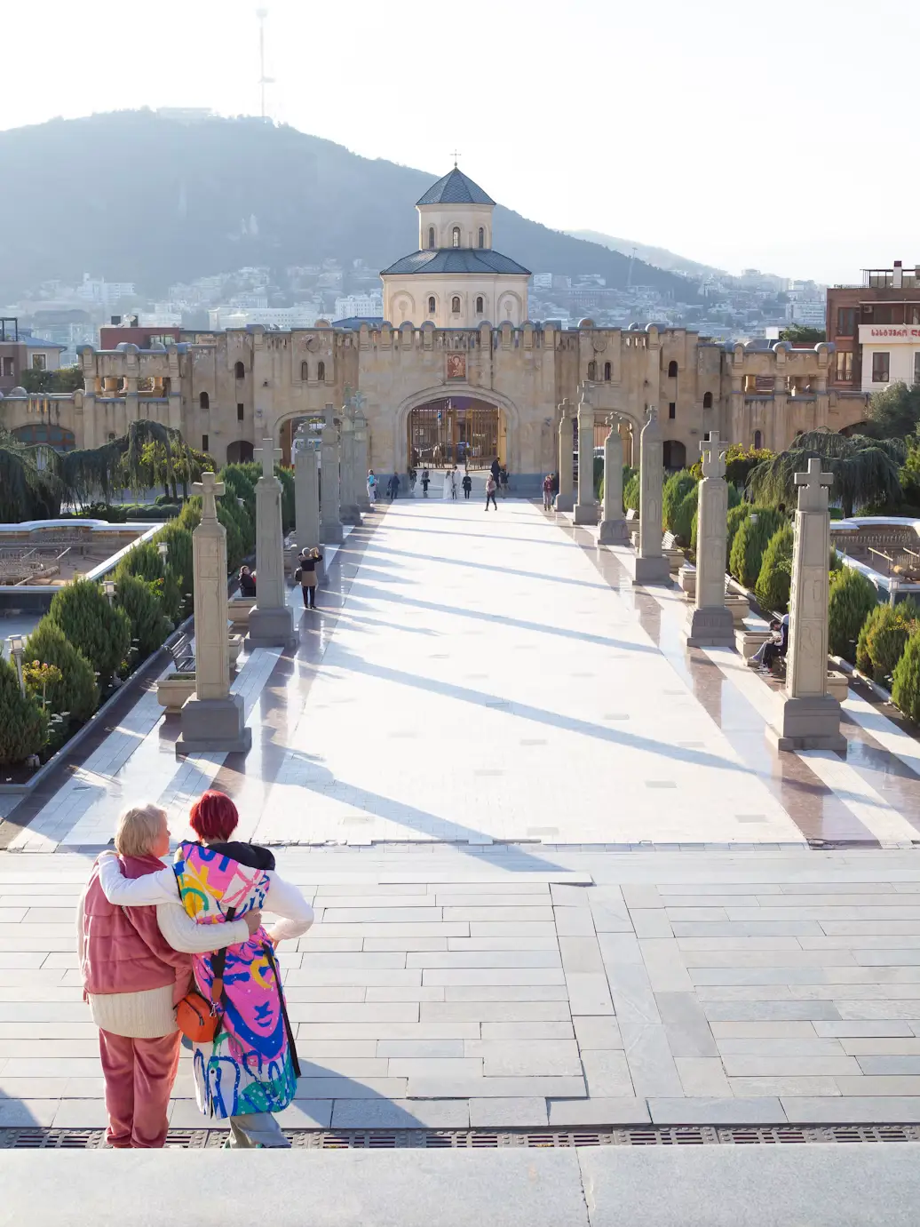 Two people embracing while overlooking a grand cathedral entrance and tree-lined walkway with hills in the background.