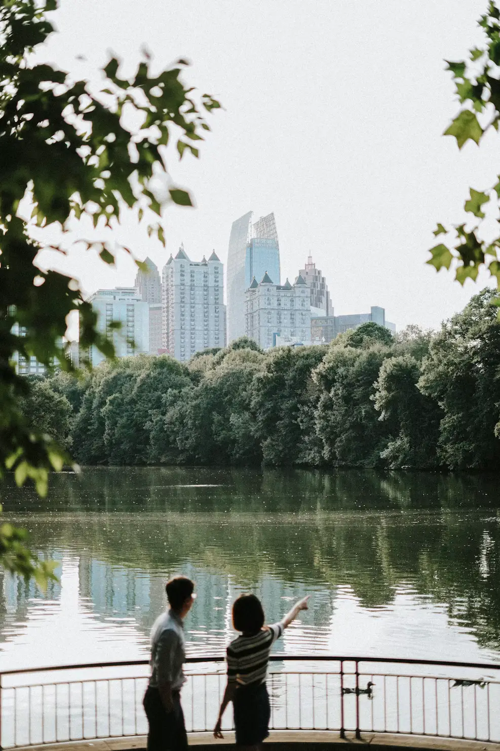 Two people stand by a lakeside railing, looking toward a city skyline beyond the trees.