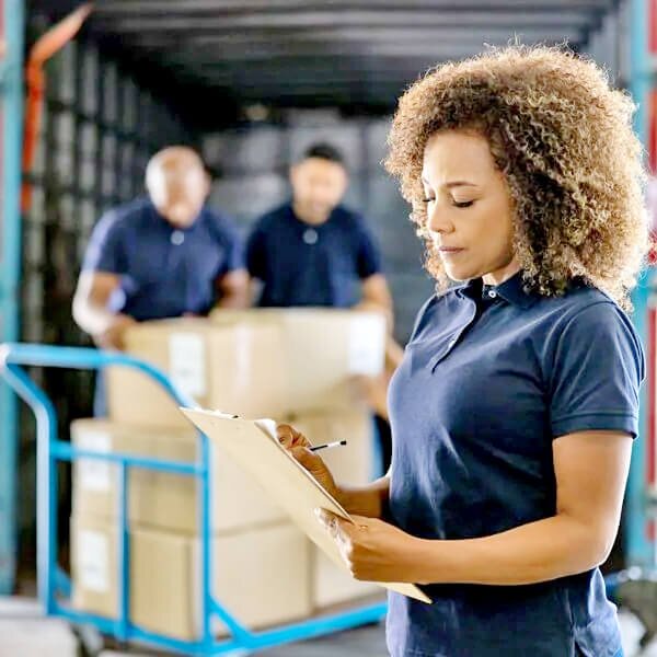 woman writing on clip board checking order of delivery men behind her holding boxes