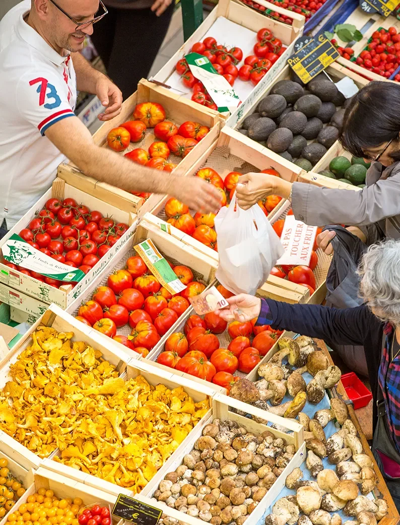 Stand de fruits et légumes colorés avec tomates, champignons et avocats. Échange entre commerçant et client sur un marché proche de Longeville-sur-Mer.