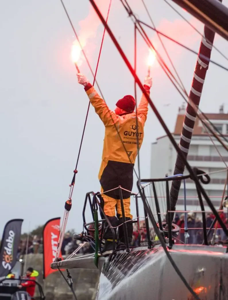 Groupe de supporters enthousiastes sur les rochers brandissant des fumigènes colorés et des pancartes près de l’eau lors d'une arrivée du Vendée Globe..