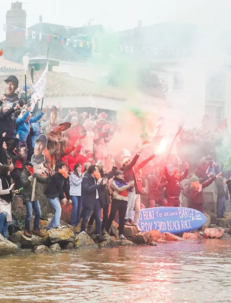 Groupe de supporters enthousiastes sur les rochers brandissant des fumigènes colorés et des pancartes près de l’eau lors d'une arrivée du Vendée Globe..