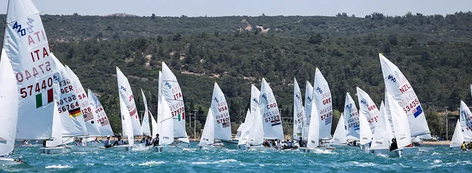 Flotte de 420 en régate sur mer turquoise avec collines en arrière-plan.