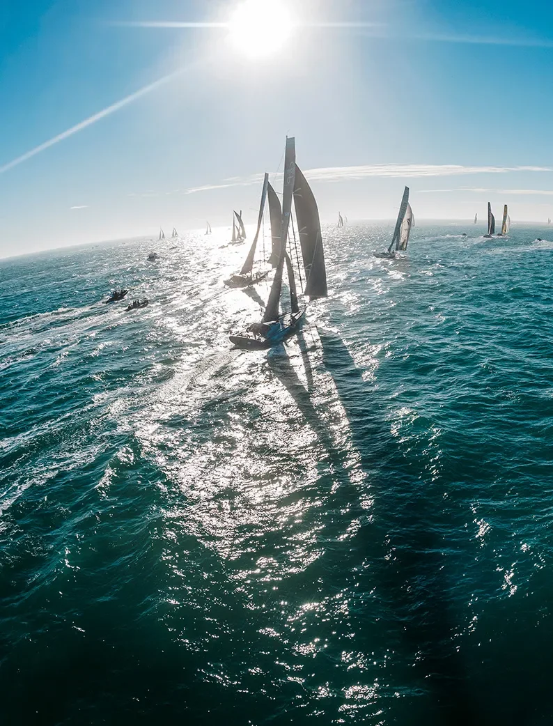 Des voiliers du Vendée Globe, aux Sables d'Olonne, naviguent sur une mer scintillante sous un ciel bleu éclatant. Le soleil brille intensément, projetant des reflets lumineux sur l'eau.