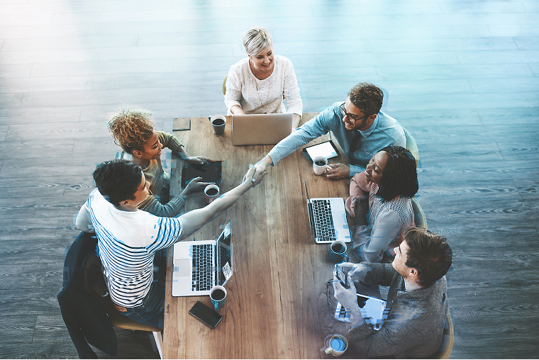 Group of people around a table with laptops, two of the people shaking hands over the table.
