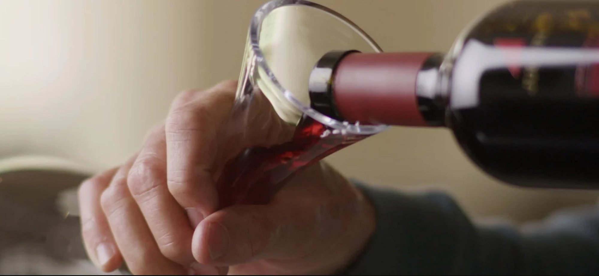 A man's hand grips a small glass, pouring a bottle of red wine into it. 