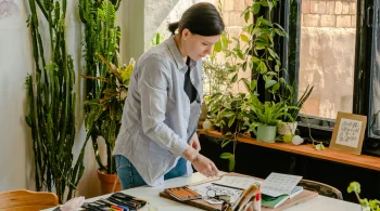 A woman stands at a desk, organizing art supplies amidst vibrant plants in a creative space