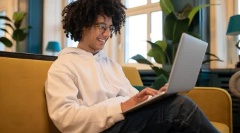 Smiling man in hoodie using laptop on a yellow sofa