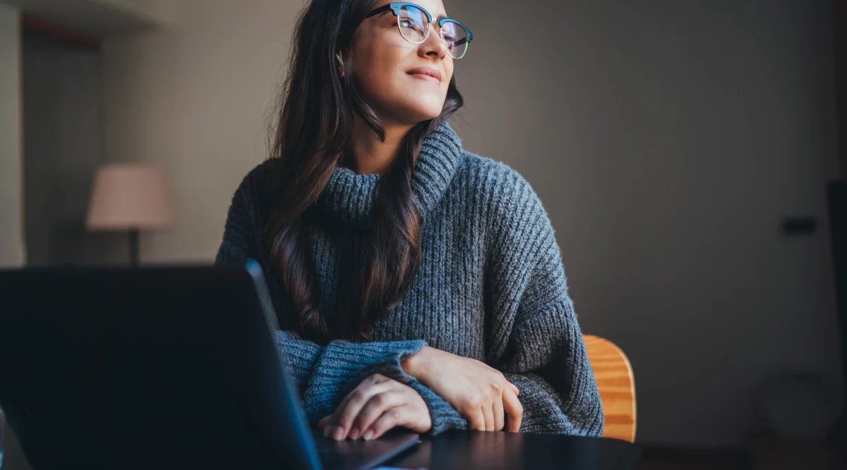 A woman sits at her desk with her laptop. She smiles and looks out the window.