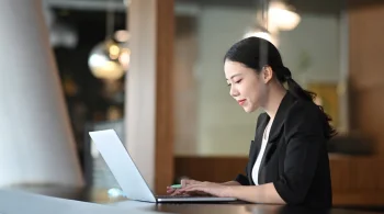 Woman in black blazer smiling while working on a laptop