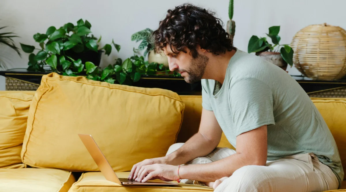 A man types on a laptop while sitting on a yellow couch surrounded by plants