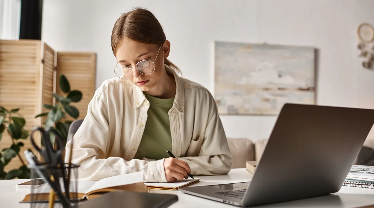 Woman writing notes at a desk with a laptop