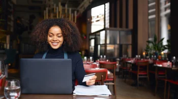 A smiling woman processes payments while working on a laptop in a lively restaurant