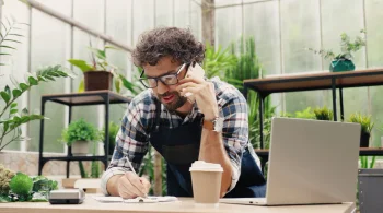 Man taking notes on a call in a greenhouse with a laptop