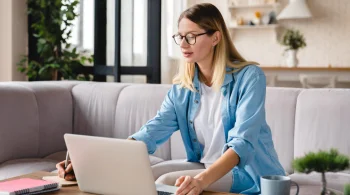 A woman in glasses sitting on the coach and working on the laptop
