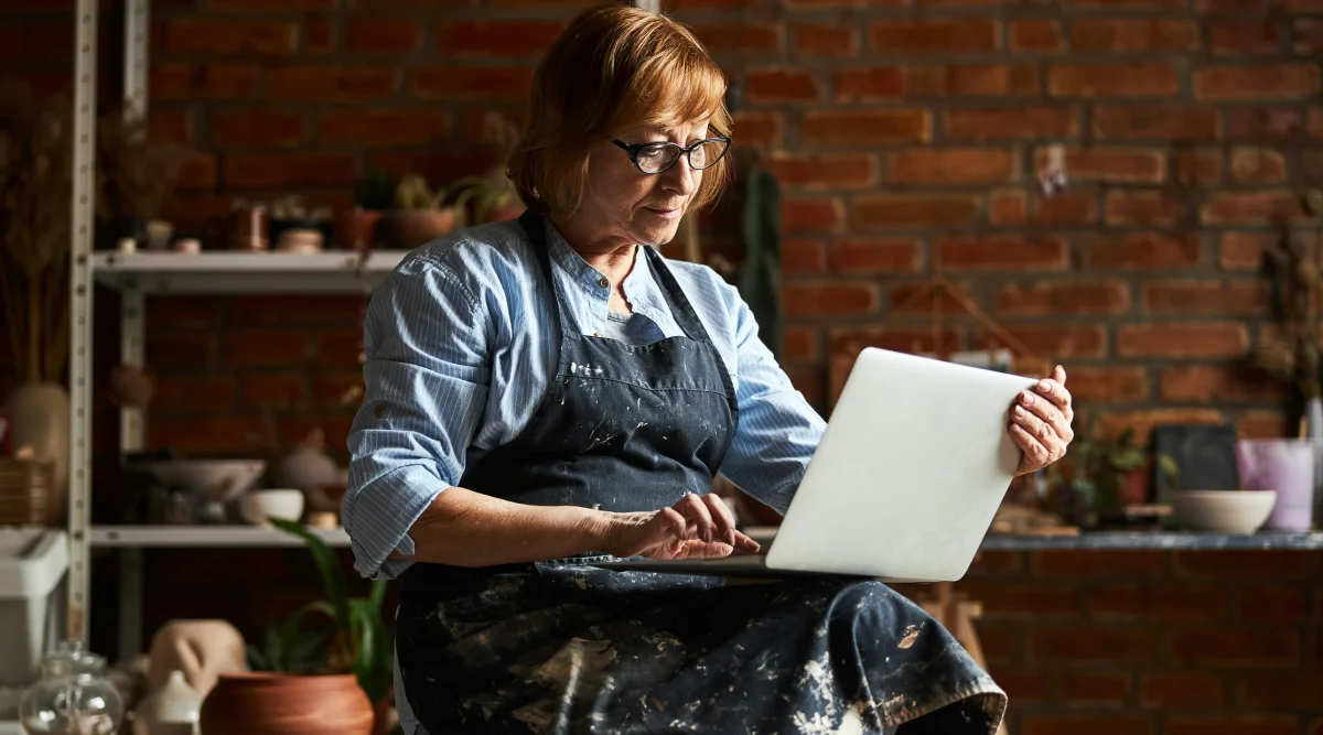 An older woman in an apron uses a laptop while sitting in a pottery studio surrounded by materials