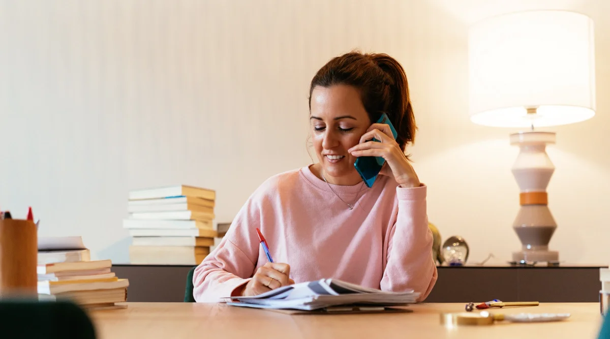 A woman in a ponytail and pink crewneck talks on the phone and takes notes while sitting at her desk.