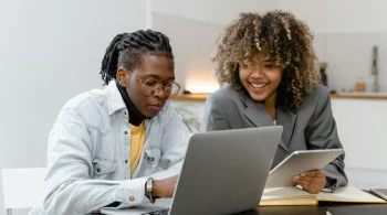 Two friends smile while collaborating on a laptop, with one holding a tablet in a cozy workspace