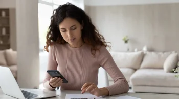 A woman reviews receipts on her phone and adds up items for an expense report using a calculator.