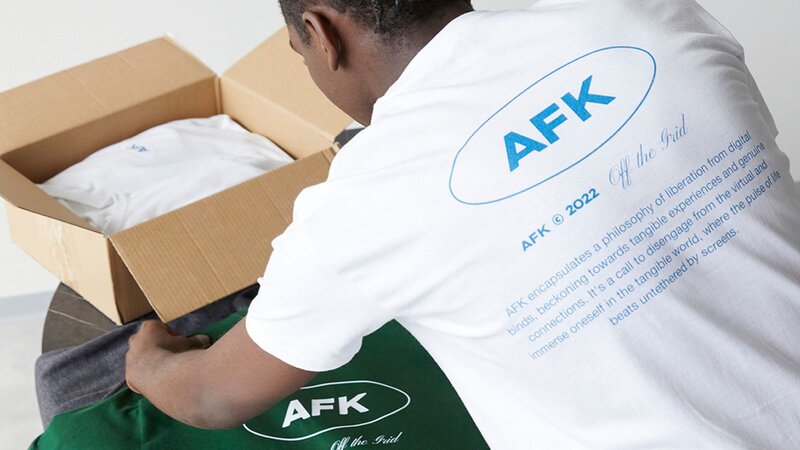 A man in a white “AFK” t-shirt is putting different-colored t-shirts on a table.