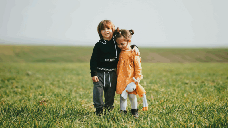 Two children standing together in a green field, smiling and enjoying a sunny day outdoors.