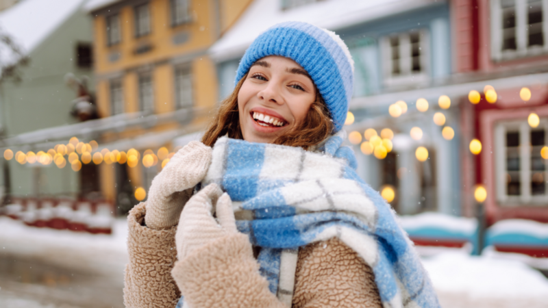 Smiling woman in winter clothes stands in a snowy street, wearing a blue hat and checkered scarf. Festive lights and houses in the background.